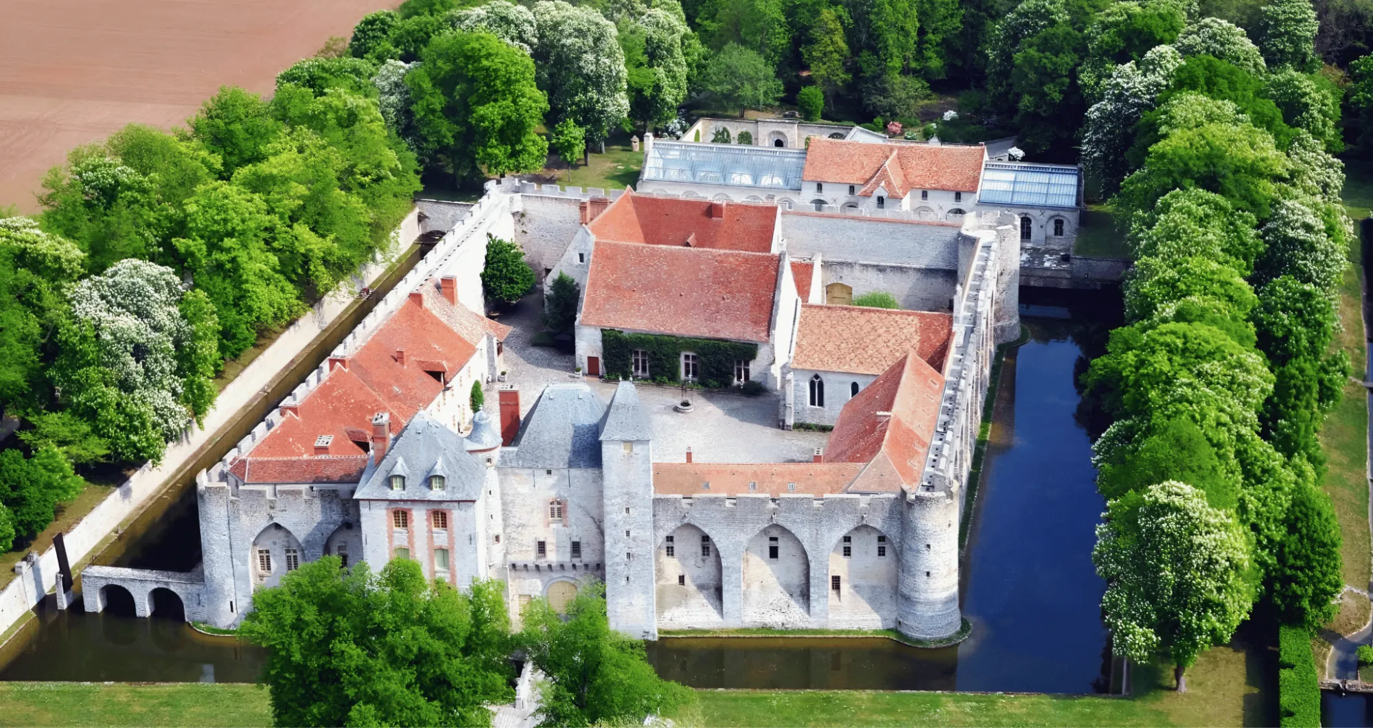 The château in its moat from the air: the courtyard, the roofs, the pool’s glass roofs behind