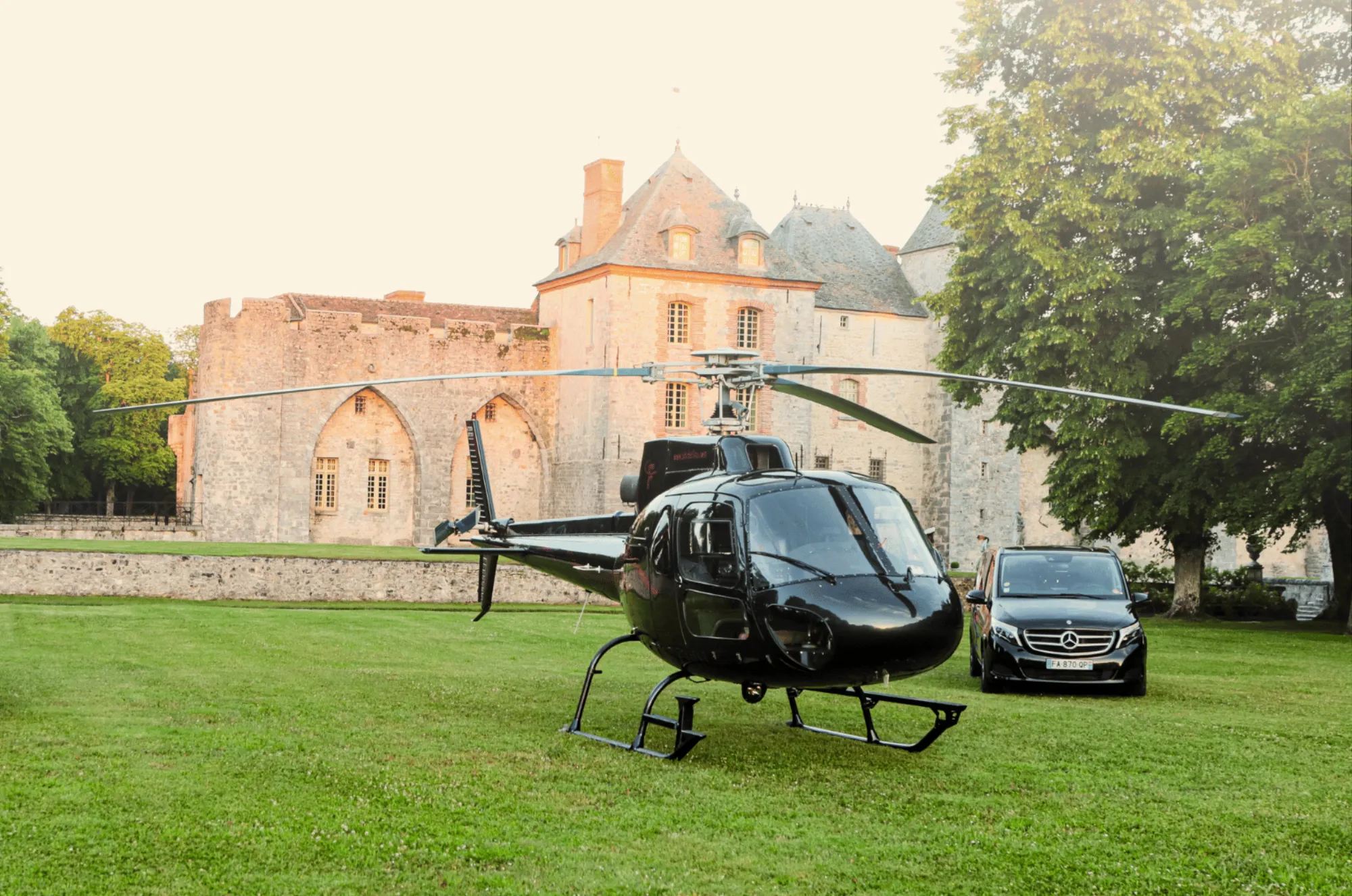 A helicopter and two chauffeured cars on the lawn, the château in evening sun